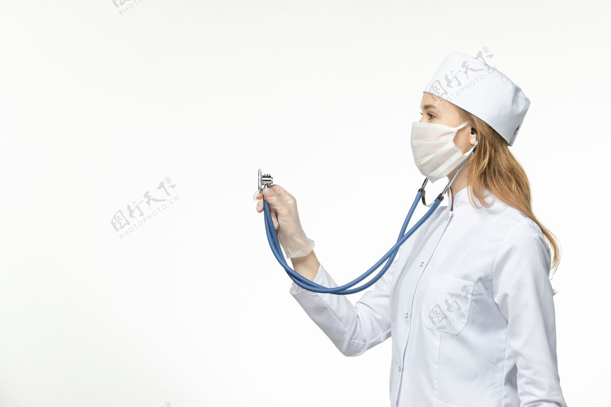 医疗正面图年轻女医生因冠状病毒穿医疗服戴口罩在白色表面使用听诊器使用专业医生