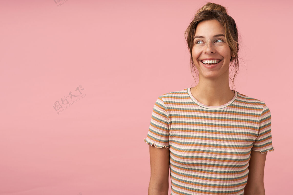 条纹年轻迷人的黑发女性的肖像画 自然的妆容 微笑着 一边愉快地看着一边 站在粉色背景下 双手放在一边情绪T恤结