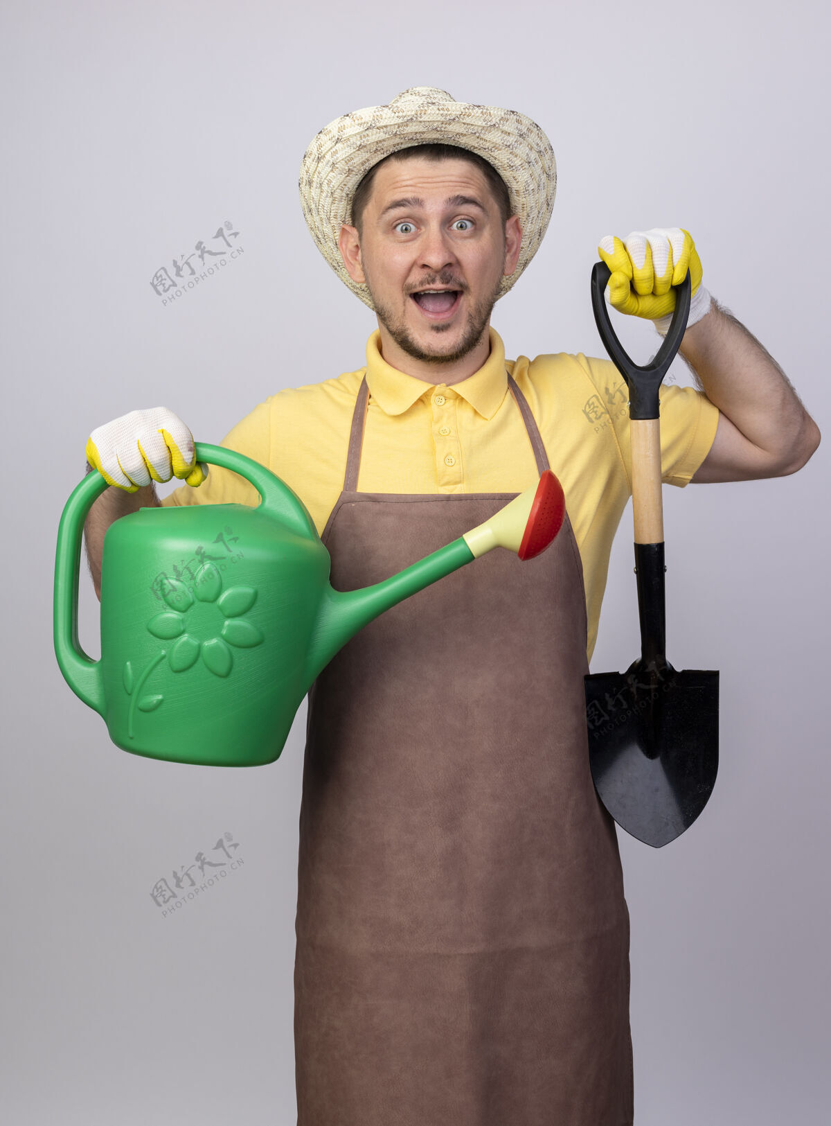 脸快乐的年轻园丁 穿着连体衣 戴着帽子 戴着工作手套 手里拿着浇水罐和铲子 满脸笑容工作欢呼铲子