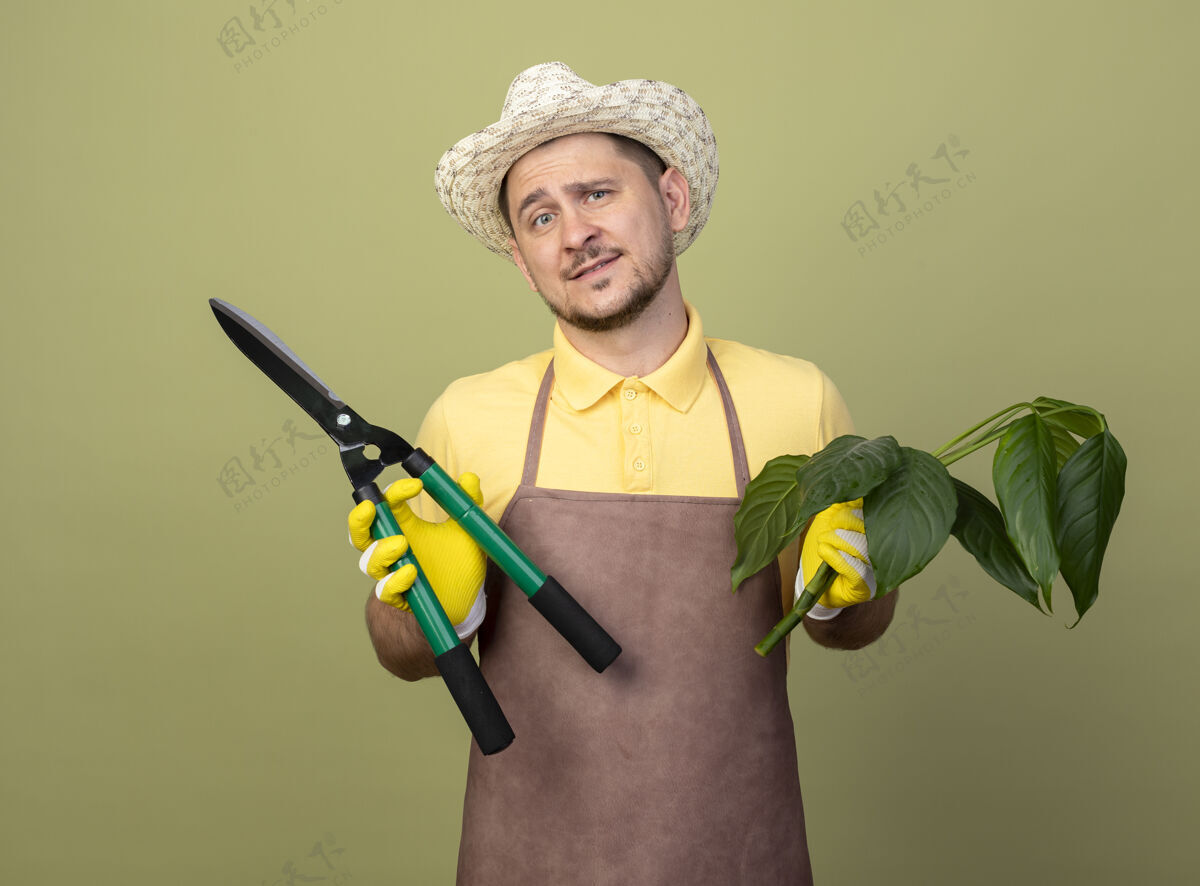 植物年轻的园丁 穿着连体衣 戴着帽子 戴着工作手套 手里拿着植物和树篱剪 满脸笑容手套微笑剪子