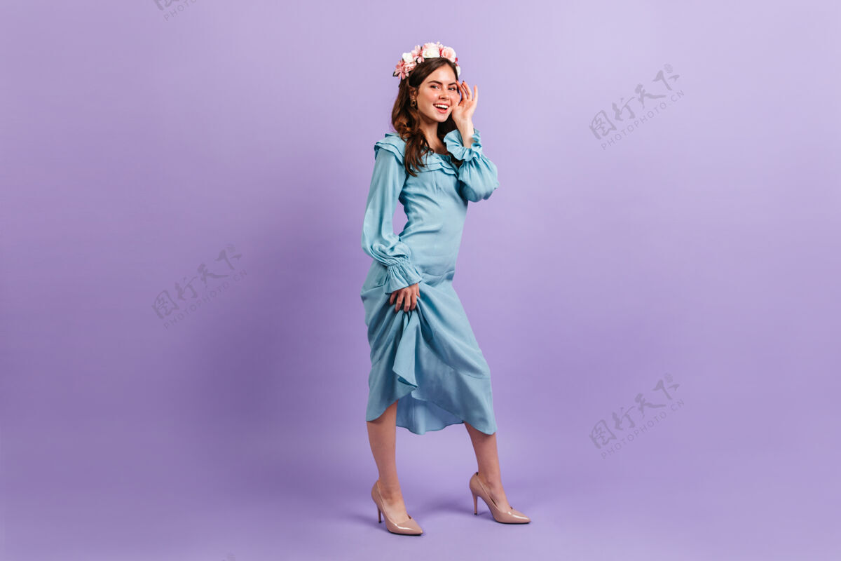 蓝色连衣裙可爱的微笑着的女士穿着缎蓝色的衣服女人在淡紫色的墙上抚摸她的脸玫瑰脸卷发