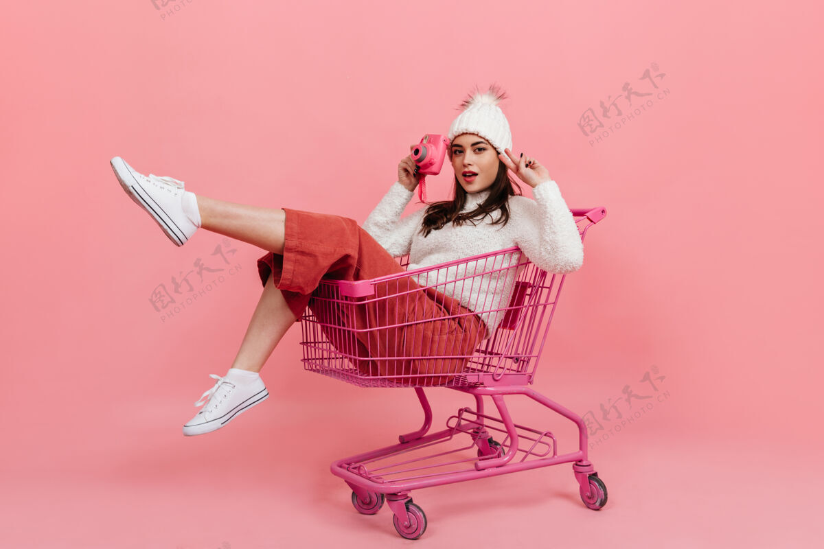 成人一个穿着针织衣服的年轻女孩坐在超市的手推车里 对着粉色的相机摆姿势波浪发模特时尚