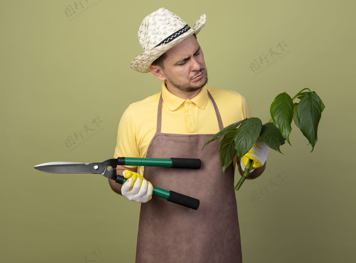 帽子年轻的园丁穿着连体衣 戴着帽子 戴着工作手套 手里拿着植物和树篱剪 表情严肃拿着工作剪子
