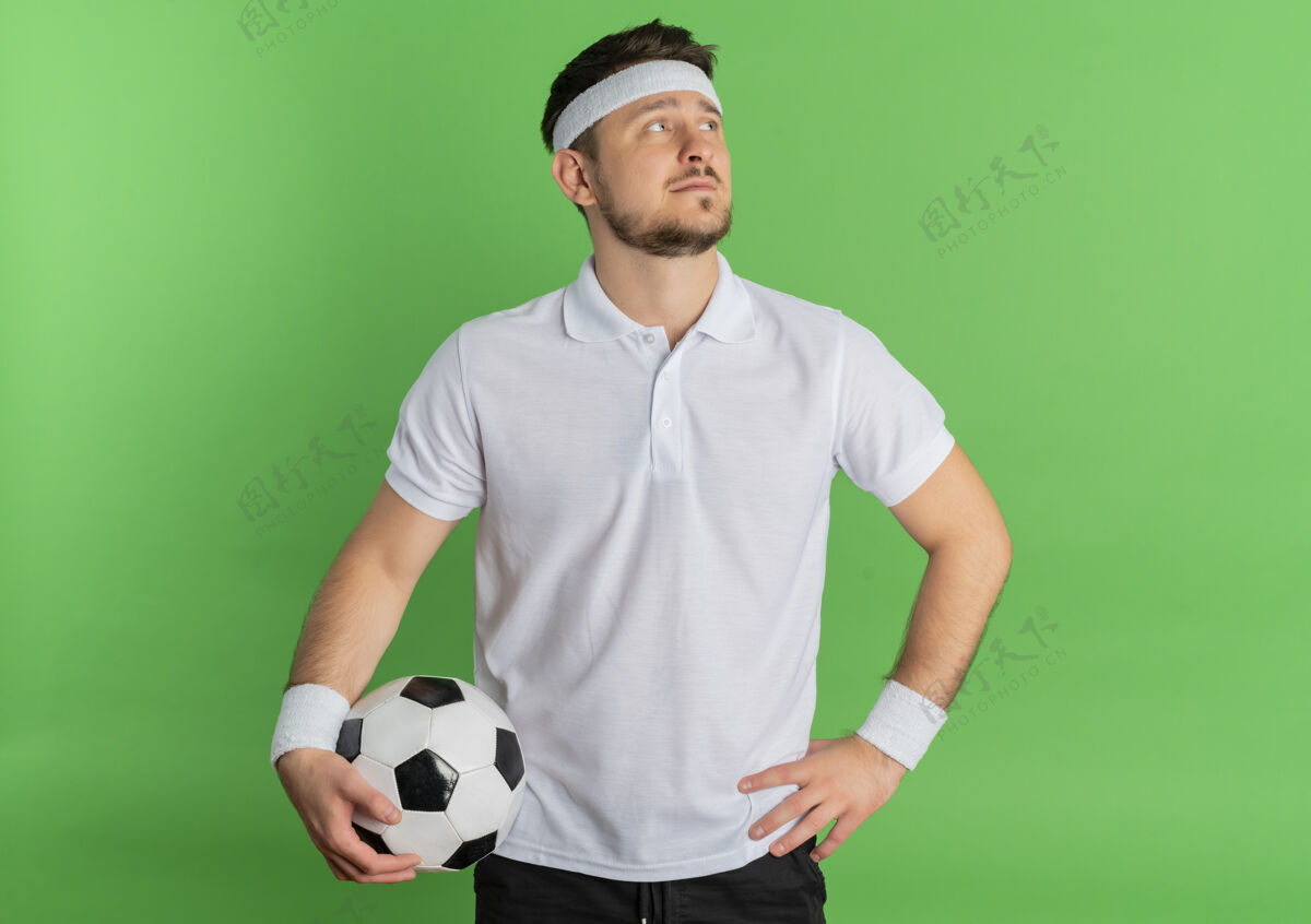 拿着身穿白衬衫 头箍 手持足球 严肃的面孔站在绿色背景下的年轻健身男子脸白色站着