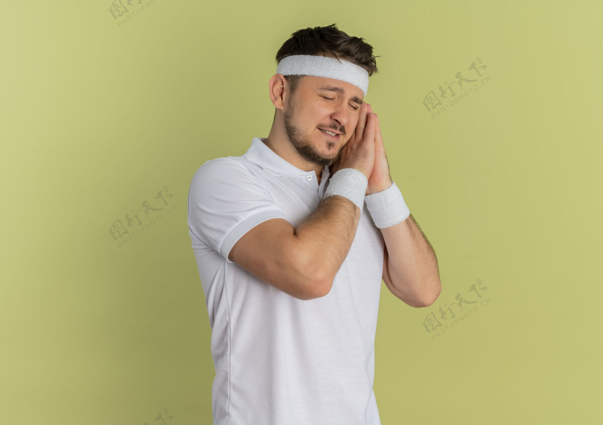 睡眠身穿白衬衫 头箍 双手合十 头靠手心 闭着眼睛的年轻健身男子想站在橄榄色背景上睡觉男人白色头