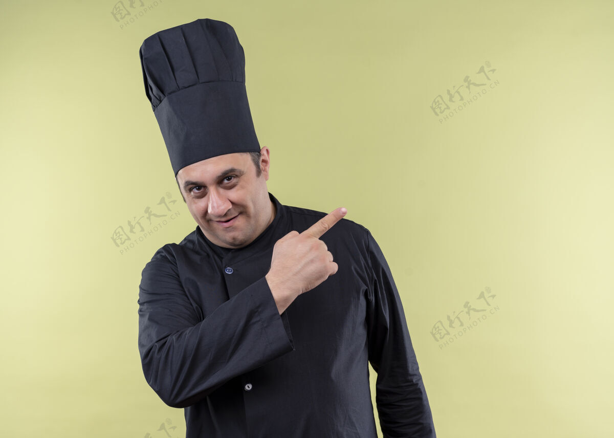 穿男厨师厨师身穿黑色制服 头戴厨师帽 看着摄像机 微笑着 用食指指着站在绿色背景上的一边看微笑指向