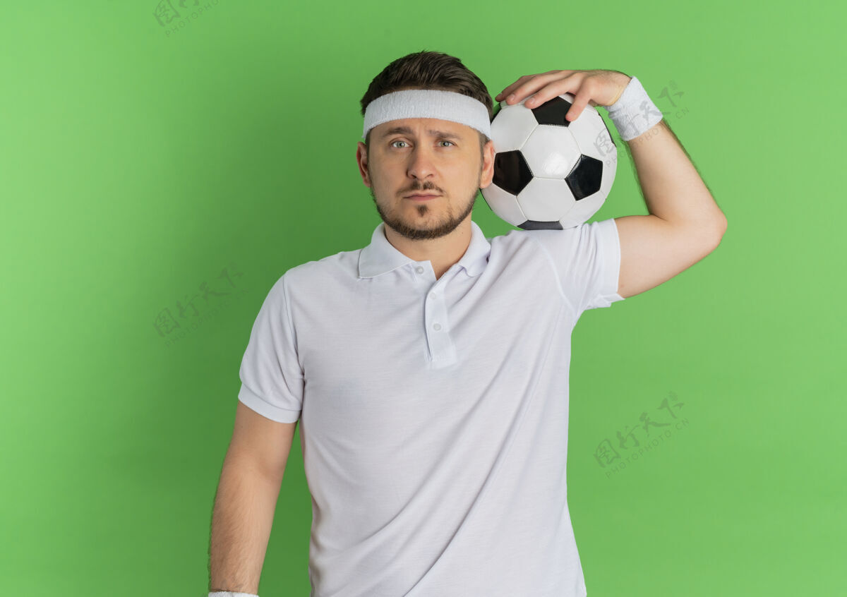 衬衫身穿白衬衫 头箍 肩上扛着足球的年轻健身男子站在绿色背景下严肃地看着摄像机站着头带健身