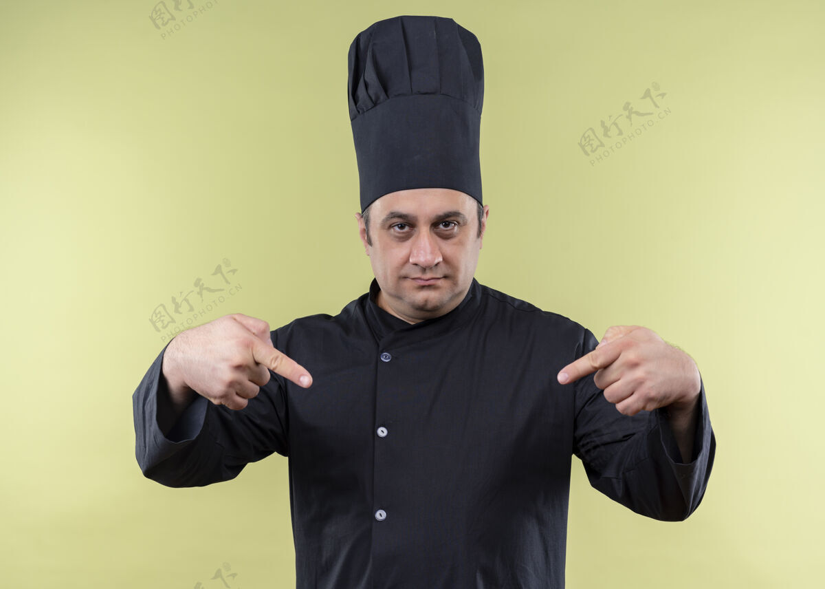 手指男厨师身穿黑色制服 头戴厨师帽 自信地用手指着站在绿色背景下的自己手指帽子烹饪