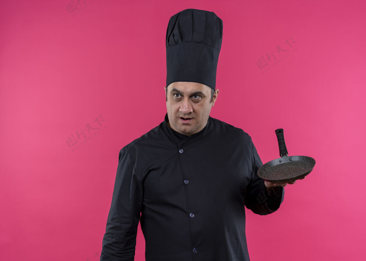 粉色男厨师身穿黑色制服 头戴厨师帽 手拿平底锅 站在粉色背景下惊讶地向一旁望去制服穿拿着