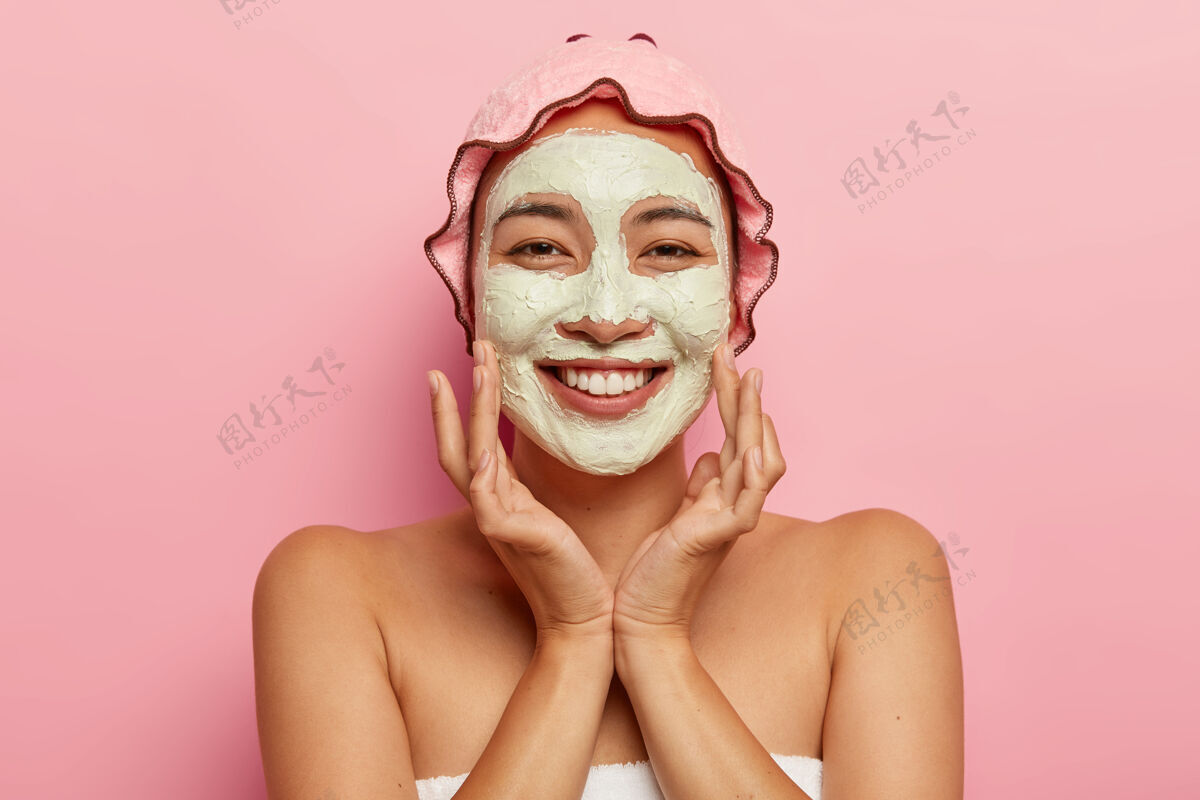 美容师适合所有年龄段的皮肤护理快乐的亚洲女士 脸上涂着去皮泥面膜 做美容 看起来很愉快 摸脸颊 戴着浴帽快乐按摩面膜