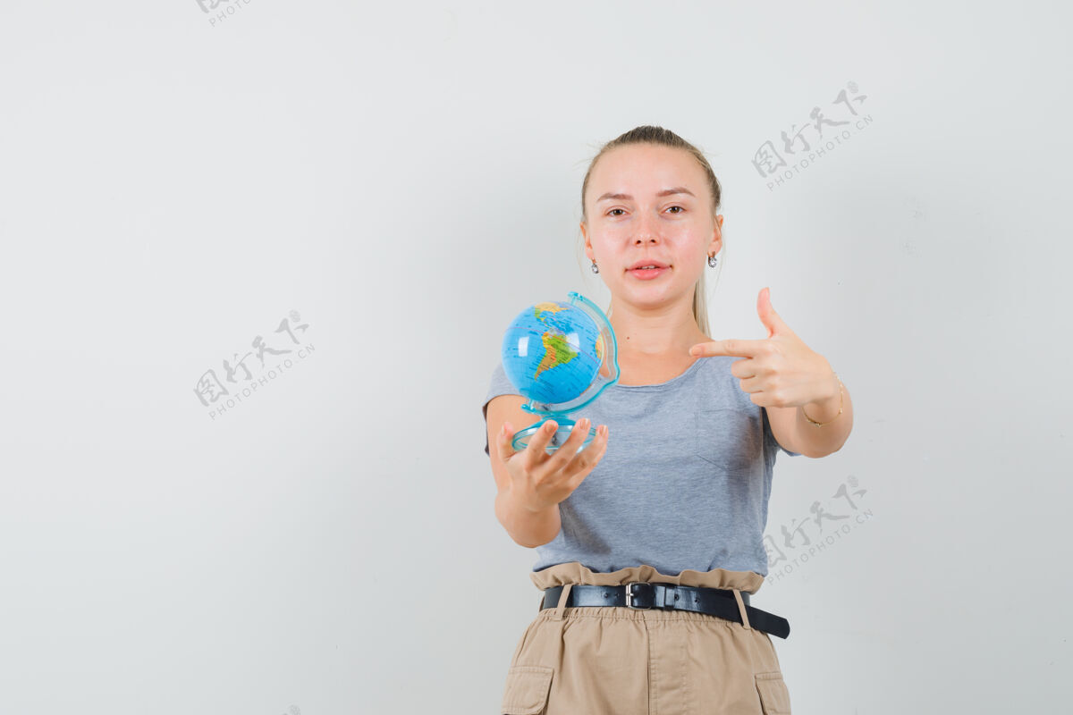 漂亮年轻的女性穿着t恤 裤子指着地球模型和微笑 前视图年轻肖像可爱
