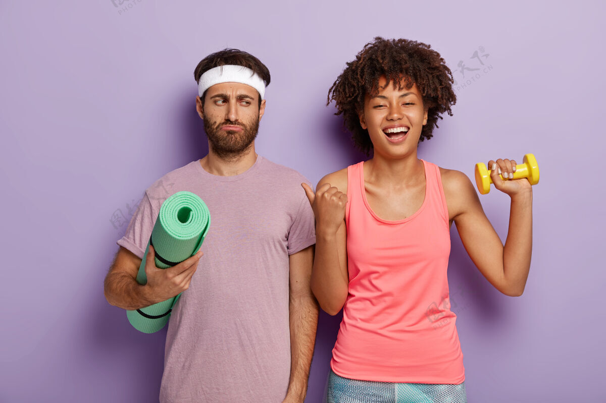 哑铃高兴的黑人发型美女用拇指指着表情不满的丈夫 一起积极锻炼 穿运动服心烦意乱的家伙不急于进行健身训练美国快乐紫色