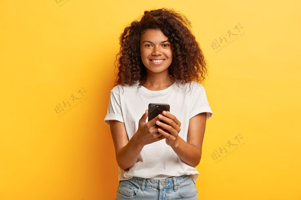 理发快乐微笑的女人手持手机 手机短信 上网 卷曲浓密的发型 穿着白色休闲t恤和牛仔裤牛仔裤使用女人