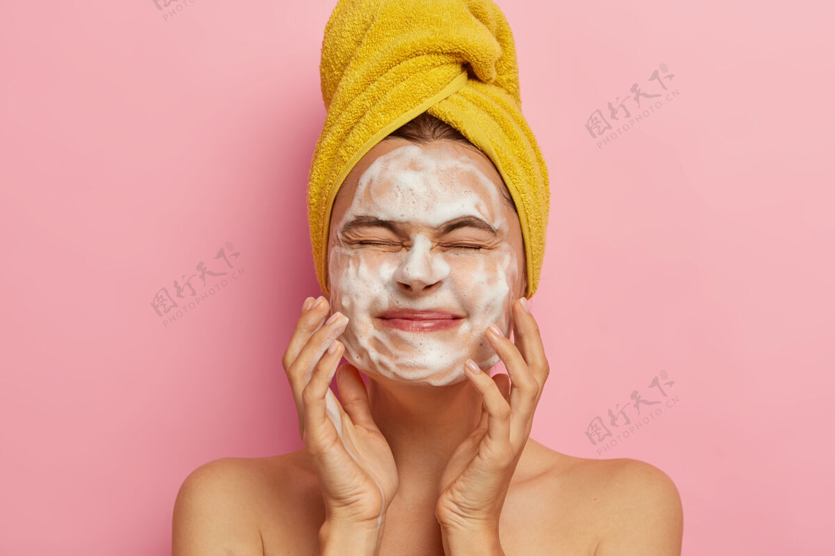 年轻快乐的年轻女性用肥皂接触面部皮肤 闭上眼睛 早上洗脸 赤裸着站在粉色的墙壁上 进行spa治疗清洁概念裸露肖像高兴