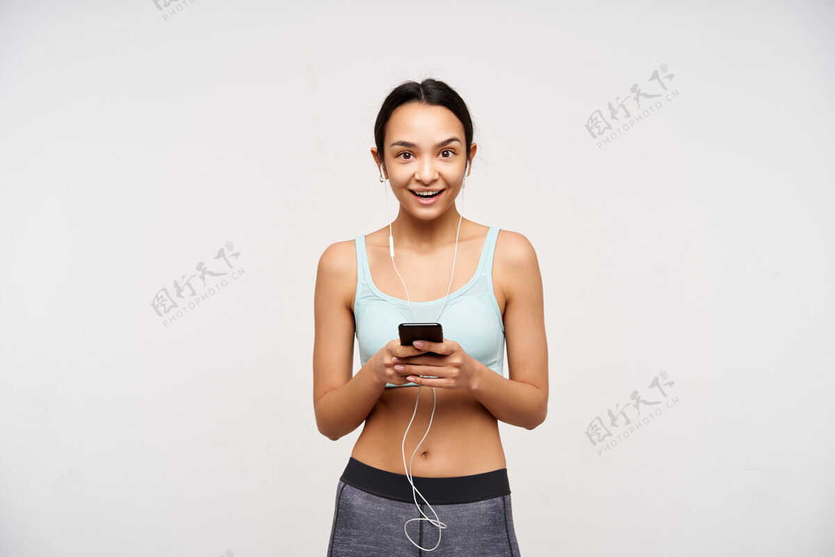 健身室内拍摄的是一个年轻的棕色眼睛 深色头发 运动型的女人举起手拿着智能手机 快乐地看着前面 隔着白墙室内化妆发型