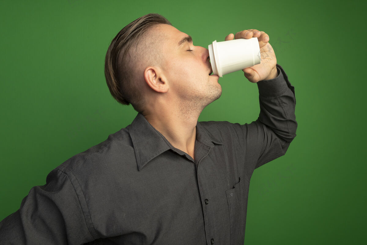 年轻穿着灰色衬衫的年轻帅哥拿着纸杯喝着咖啡站在绿色的墙上表情咖啡帅哥