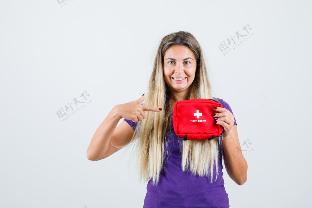 看金发女士指着急救箱在紫罗兰色的t恤衫 看起来很高兴 前视图工具包欢快美丽