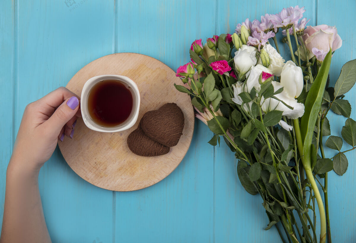 花顶视图女性手拿着一杯茶在一个木制的厨房板与美妙的鲜花隔离在一个蓝色的木制背景手景观杯子