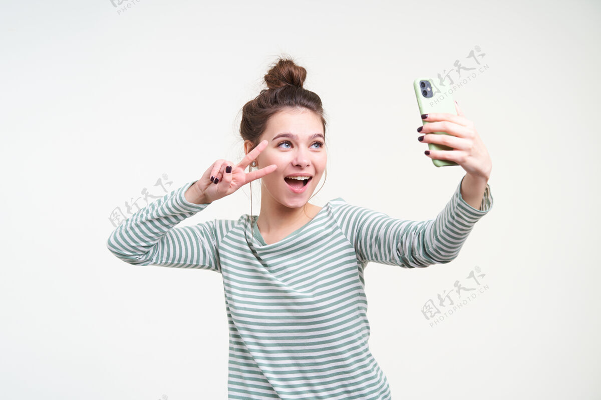 发型开朗的年轻蓝眼睛可爱的深色头发女性 发髻式 举手表示和平 一边用智能手机自拍 隔着白墙积极手机表情