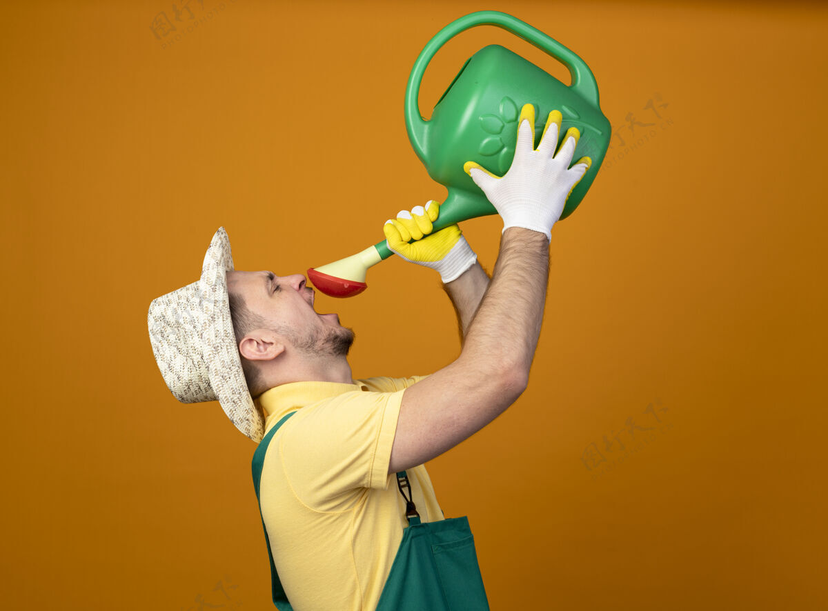 年轻人年轻的园丁穿着连体衣 戴着帽子 手里拿着浇水罐 站在橘红色的墙上 试图从中喝水帽子站人