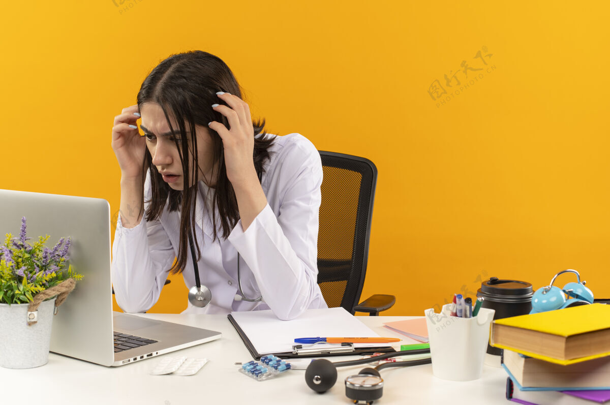 坐着身穿白大褂的年轻女医生拿着听诊器看着自己的笔记本电脑屏幕 困惑而焦急地坐在桌边橘色墙上的文件外套人桌子