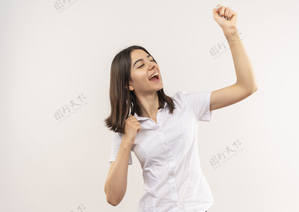 女性身穿白衬衫的年轻女孩握紧拳头 为自己的成功而欢欣鼓舞站在白墙上人人手势