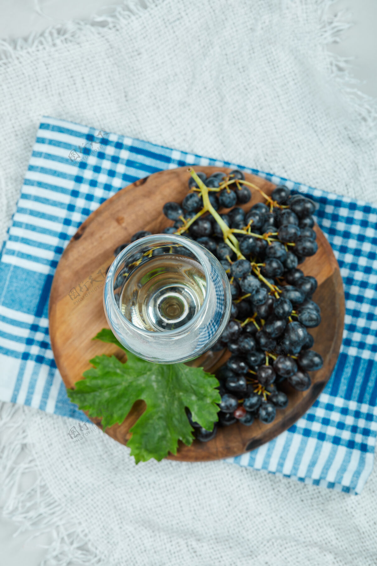 新鲜的一簇带叶子的黑葡萄和一杯白葡萄酒摆在蓝色桌布上葡萄叶子玻璃
