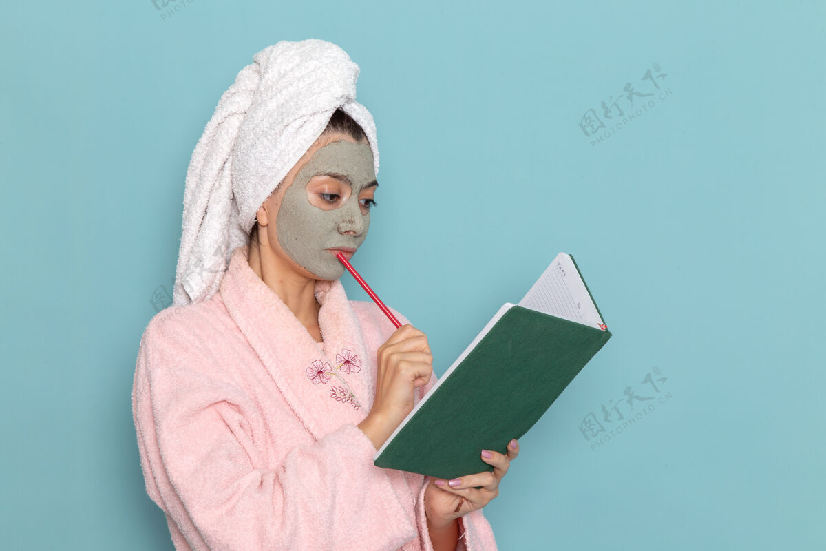 浴袍前视图穿着粉色浴袍的年轻女性在淋浴后阅读蓝色墙壁上的抄写本美容水霜自我护理淋浴浴室后成人自理
