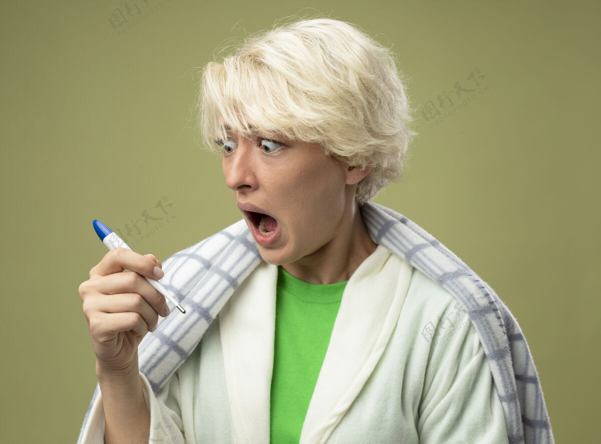 包裹生病的不健康的女人 短发裹在毯子里 看着温度计 不舒服 站在光墙上 感到震惊不适震惊毯子