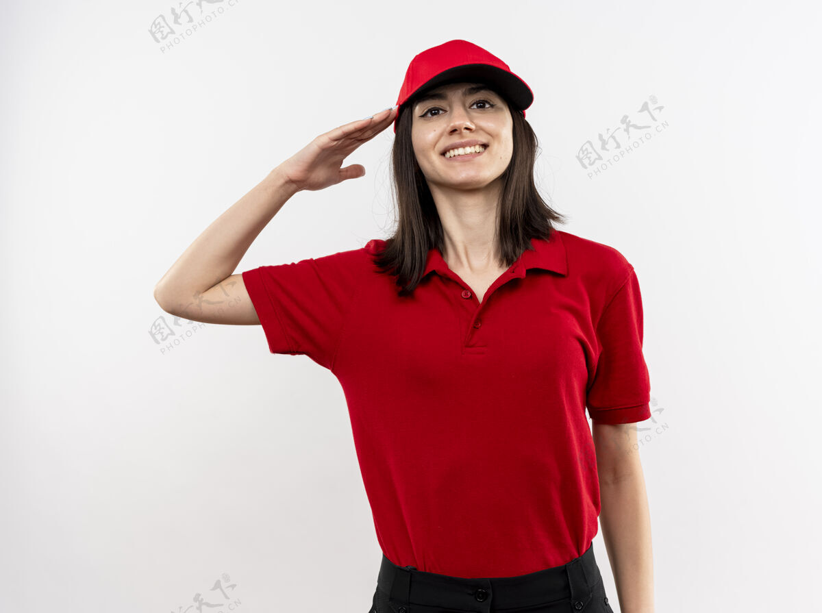 年轻年轻的送货员身穿红色制服 头戴礼帽 站在白色的墙边 微笑着自信地致意递送看帽子