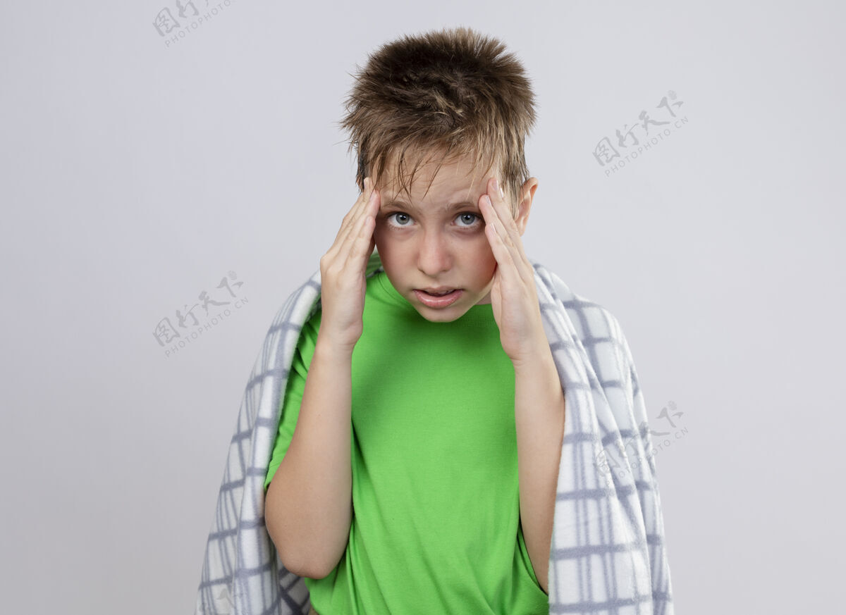 触摸生病的小男孩穿着绿色t恤 裹着毛毯 感觉不舒服 摸着太阳穴 站在白墙上感到疼痛站立男孩疾病