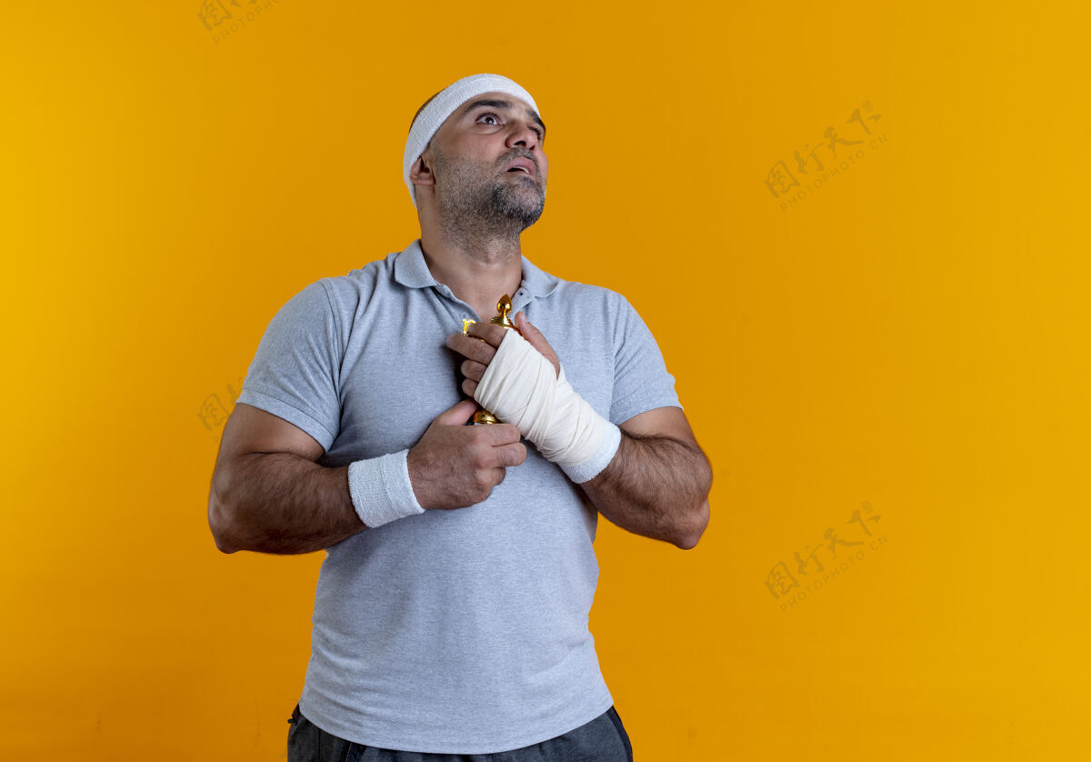 教练戴着头巾的成熟运动型男人抱着他的奖杯 站在橙色的墙上 带着希望的表情仰望着运动装运动人