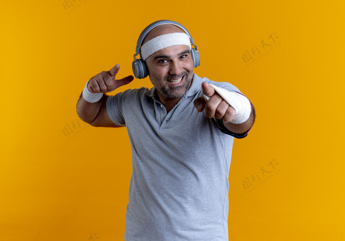 男戴着头巾的成熟运动型男人 头上戴着耳机 手指指着前面 站在橘色的墙上愉快地微笑着成熟运动教练