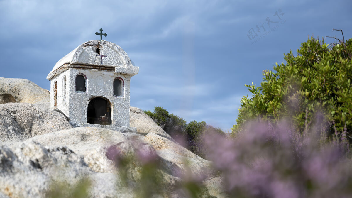 东正教一座古老而小巧的神龛坐落在爱琴海沿岸的岩石上 四周灌木丛生 天空多云 希腊纪念碑基督教十字架微型