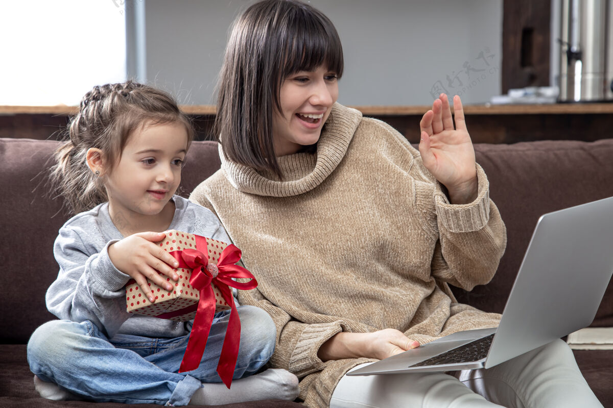 电脑快乐的一家人看着笔记本电脑屏幕打长途视频电话微笑的妈妈和带着礼盒的小女孩对着网络摄像头聊天礼物沟通娱乐