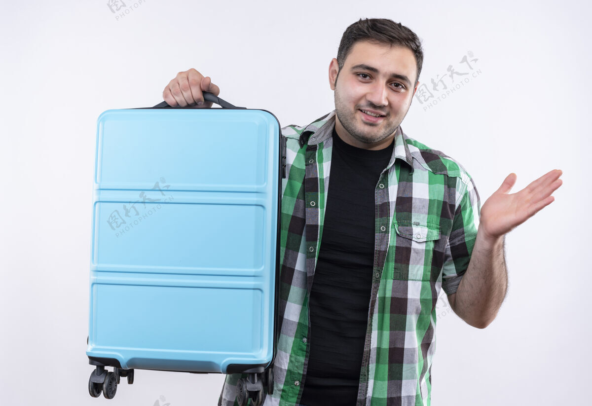 年轻人穿着格子衬衫的年轻旅行家拿着手提箱 看起来很困惑 胳膊伸向一边 微笑着站在白色的墙上困惑衬衫男人