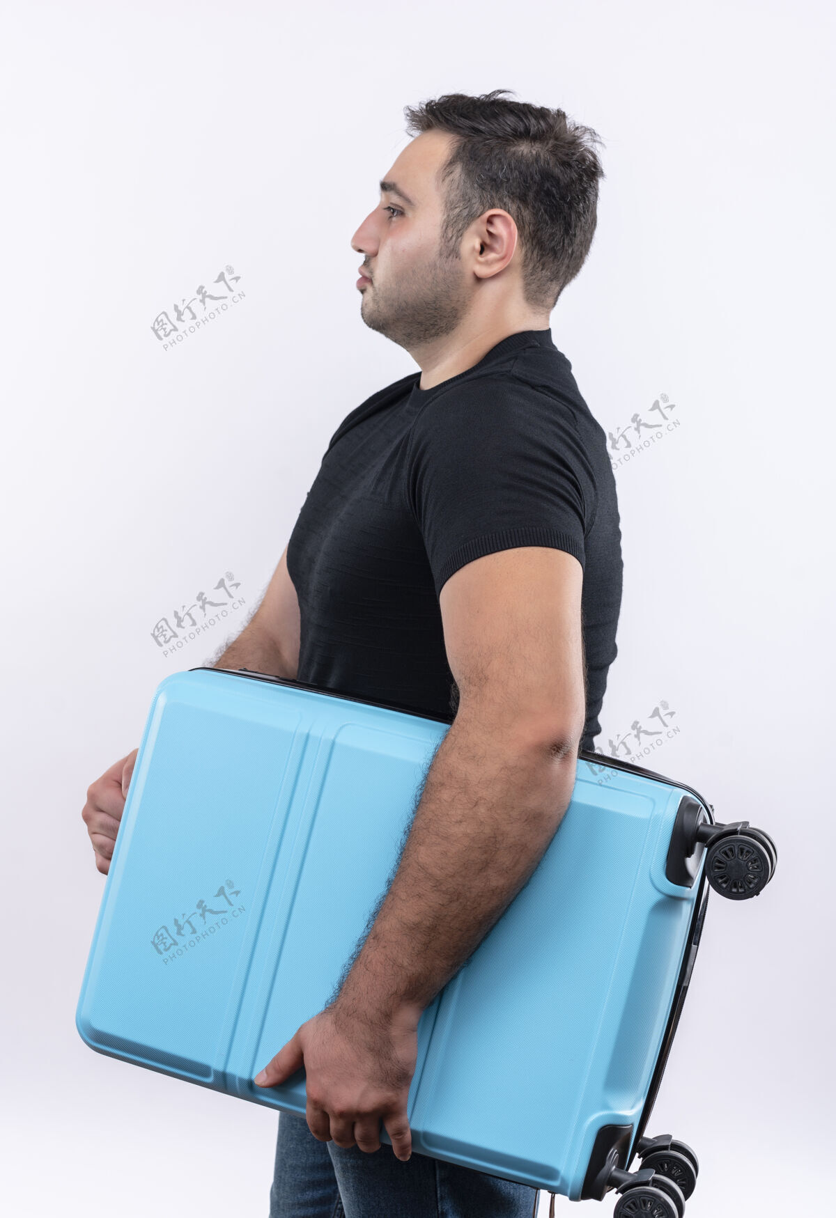 人穿着黑色t恤的年轻旅行家手拿着手提箱站在一边 严肃的脸越过白墙侧身假期手提箱