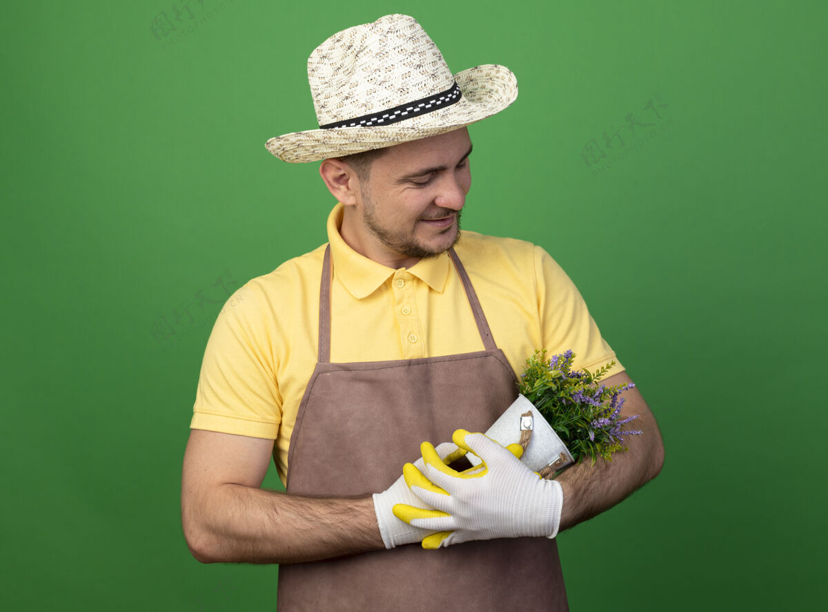 人年轻的园丁穿着连体衣 戴着帽子 戴着工作手套 手里拿着盆栽植物 站在绿色的墙上 带着爱的微笑看着它站着帽子锅