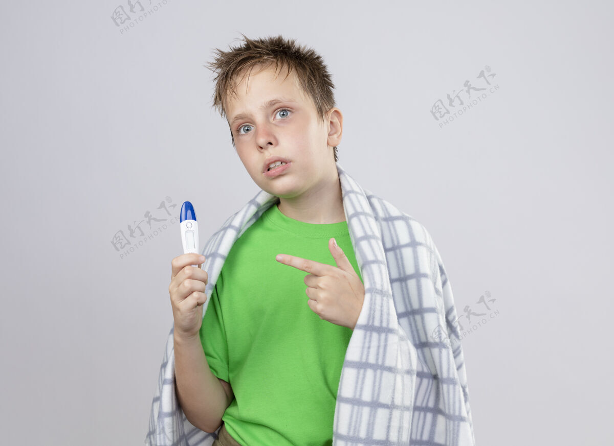 不舒服生病的小男孩穿着绿色t恤 裹着毛毯 站在白色的墙上拿着温度计 用食指指着它 感觉不舒服小感觉男孩