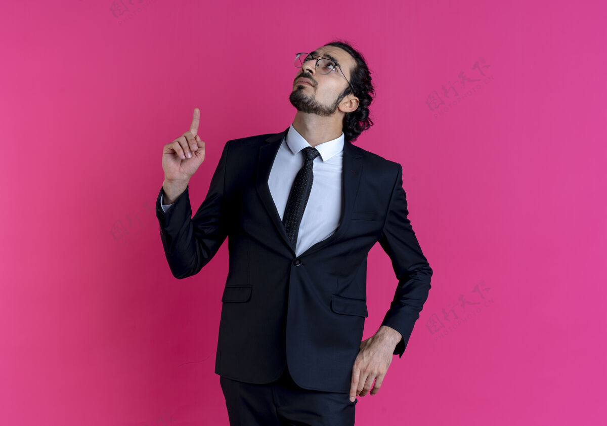 看一个穿着黑色西装 戴着眼镜 手指朝上的商人站在粉红色的墙上 看上去很困惑男男人人