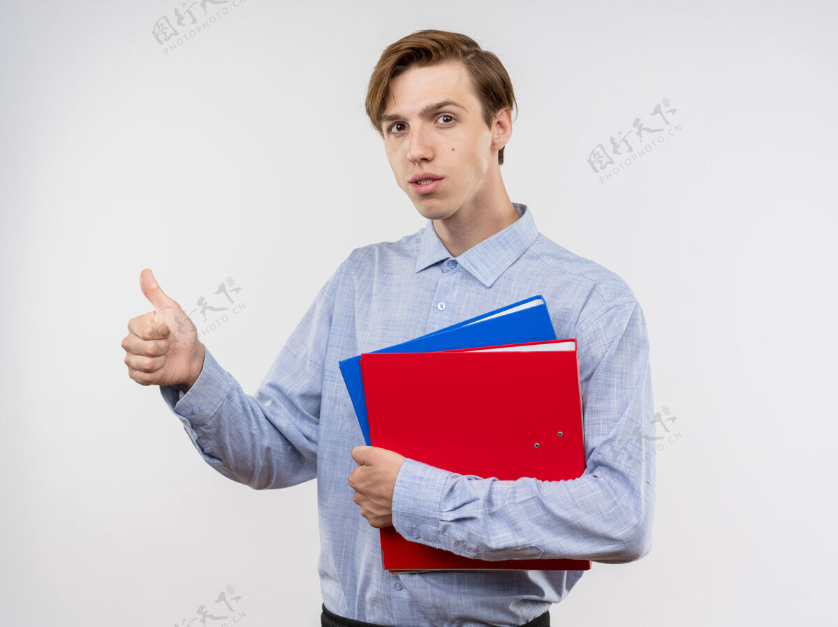 年轻穿着蓝色衬衫的年轻人拿着文件夹 竖起大拇指 自信地站在白色的墙上拇指夹着抱着
