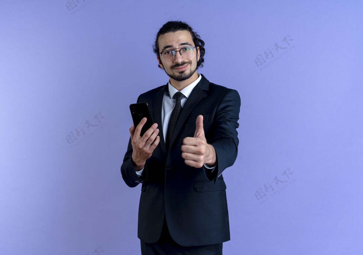 眼镜身穿黑色西装 戴着眼镜 手持智能手机的商人站在蓝色的墙上 朝前竖起大拇指微笑着微笑手机男士