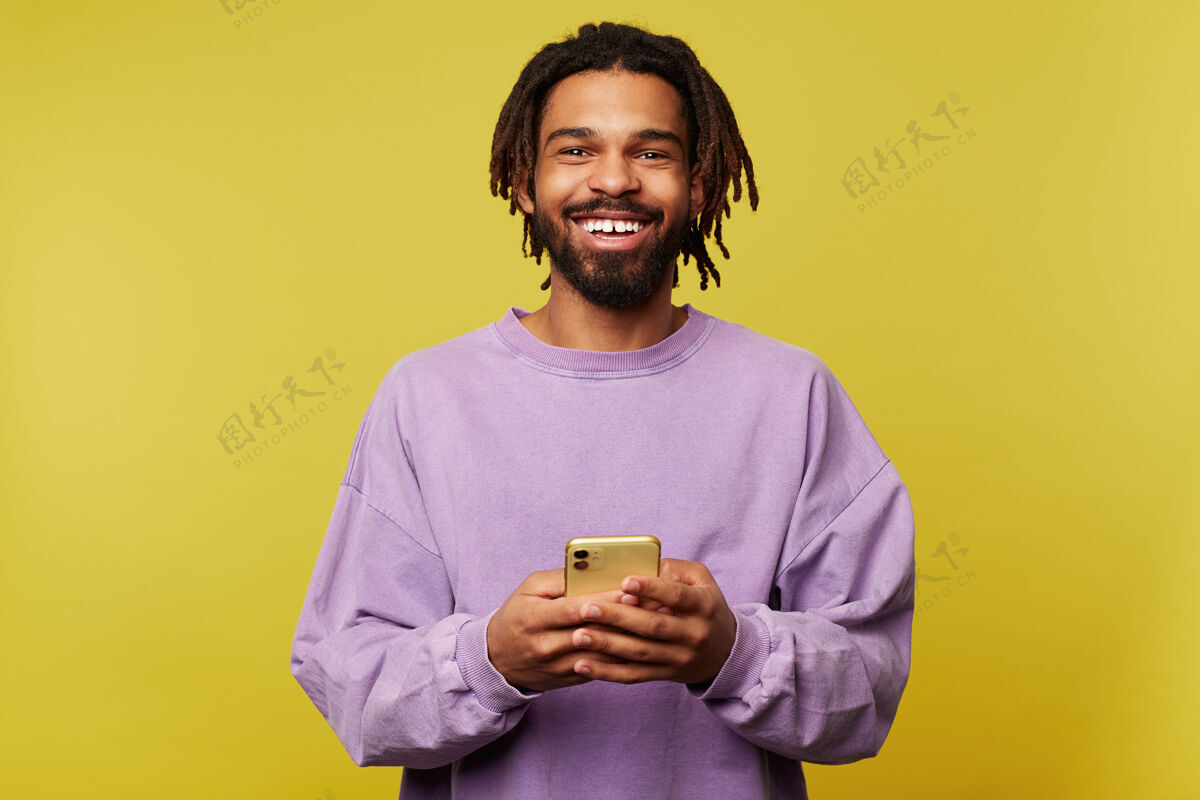 男性快乐的年轻帅气的棕色头发男人 留着可怕的长发 拿着手机对着镜头愉快地微笑 穿着紫色运动衫 在黄色背景下被隔离鬃毛胡须手机