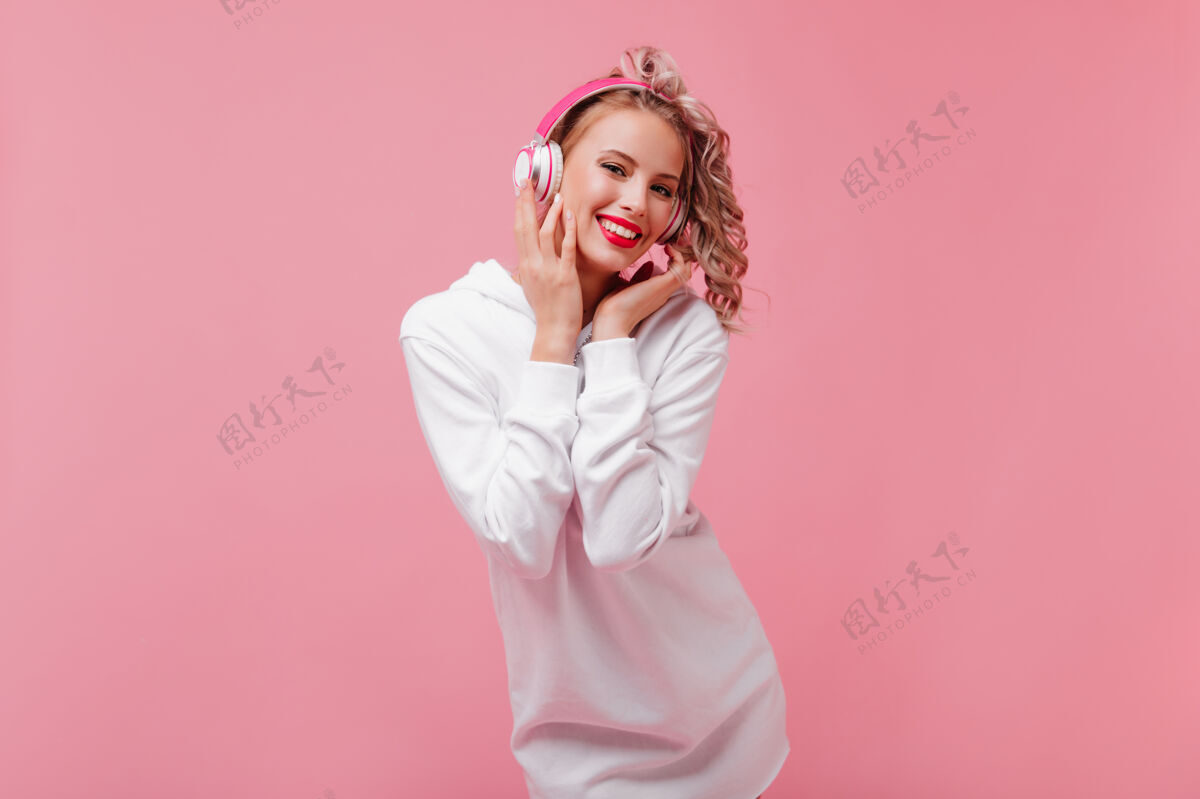 女性年轻女子摆姿势 通过她的粉红色耳机听音乐可爱夏天卷发