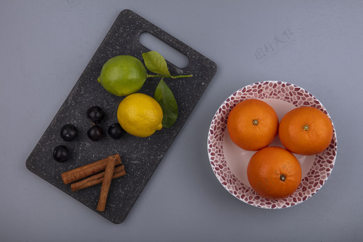 柠檬俯瞰图：柠檬 樱桃 李子和肉桂放在砧板上 橘子放在盘子里 背景是灰色的柑橘橘子切