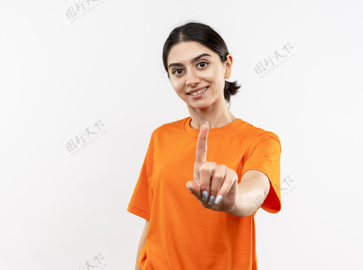 橙色穿着橙色t恤的年轻女孩站在白色的墙上 脸上露出微笑的食指年轻穿微笑