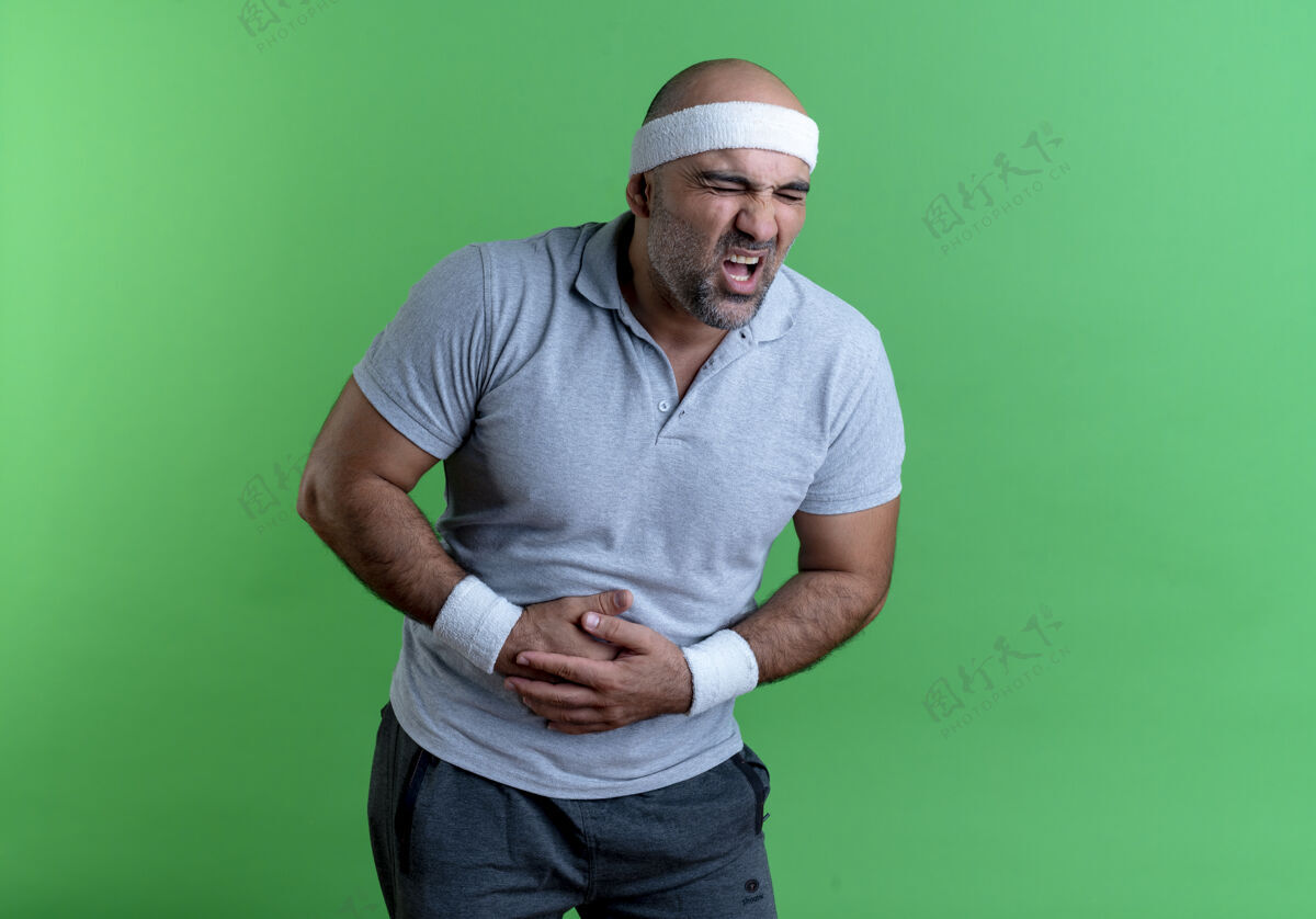 姿势戴着头巾的成熟运动型男人 看起来身体不适 站在绿墙上摸着肚子 疼痛难忍运动男人市民