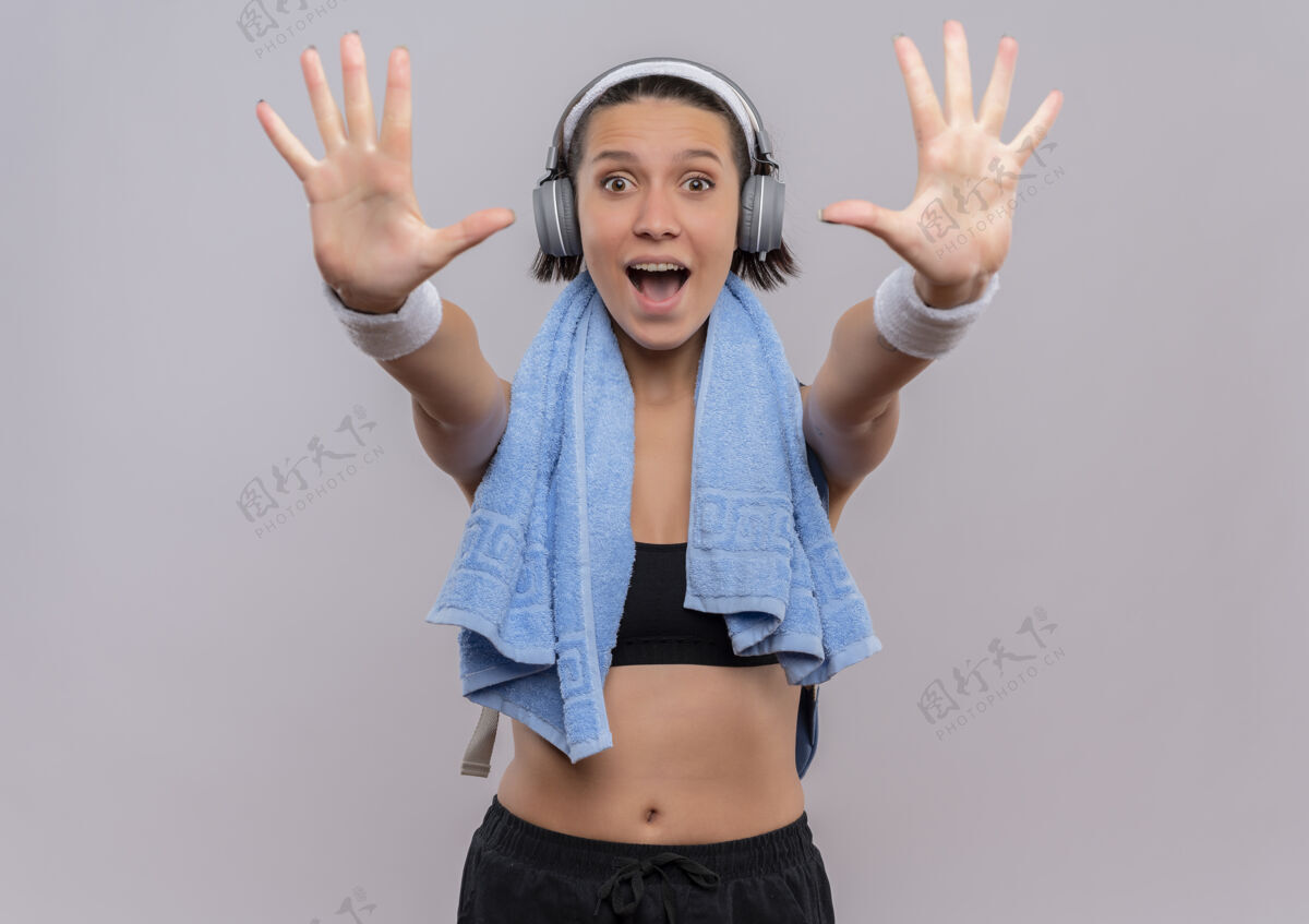 头一位身穿运动服的年轻健身女士头戴耳机 脖子上套着毛巾 伸出手掌 站在白色的墙壁上 脸上露出十号微笑 快乐和兴奋兴奋运动员女人