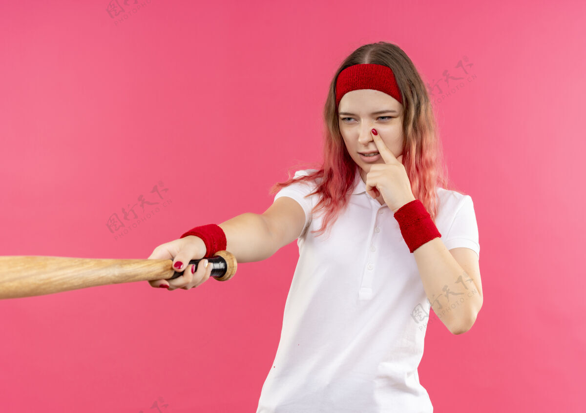 运动装戴着头巾的年轻运动型女子拿着球棒去打棒球 站在粉红色的墙上显得很自信公民看运动