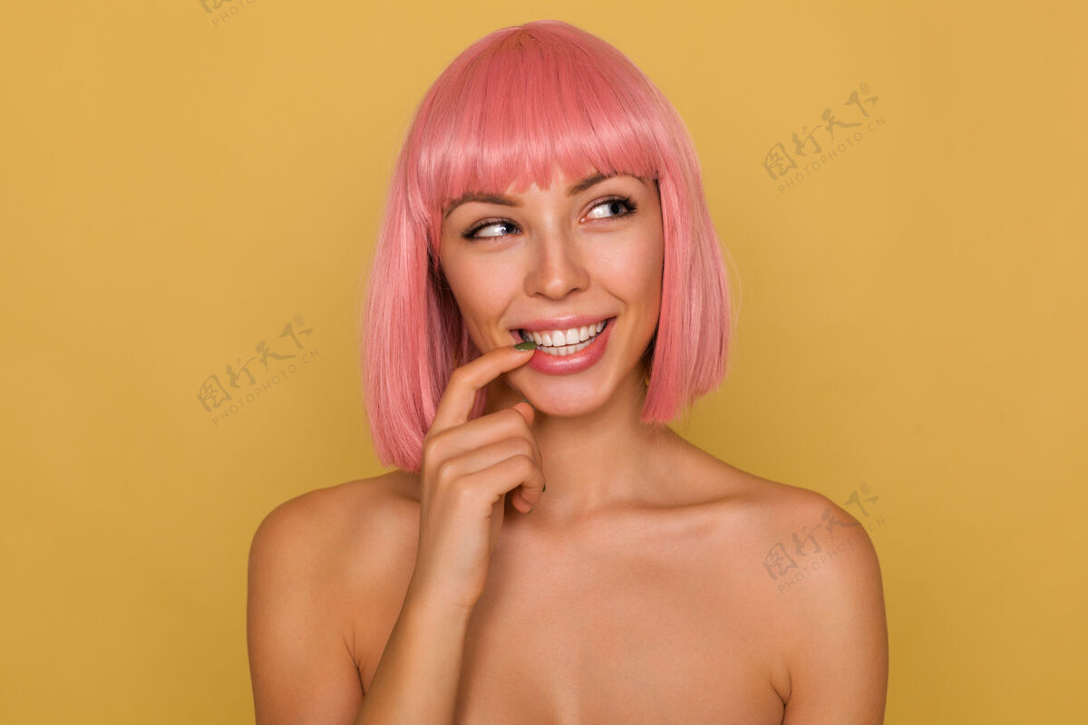 发型积极的年轻迷人的粉红色头发的女士 短发时髦 狡猾地看着旁边 微笑着用食指触摸她的内衣 在芥末墙上摆姿势20秒情感高兴